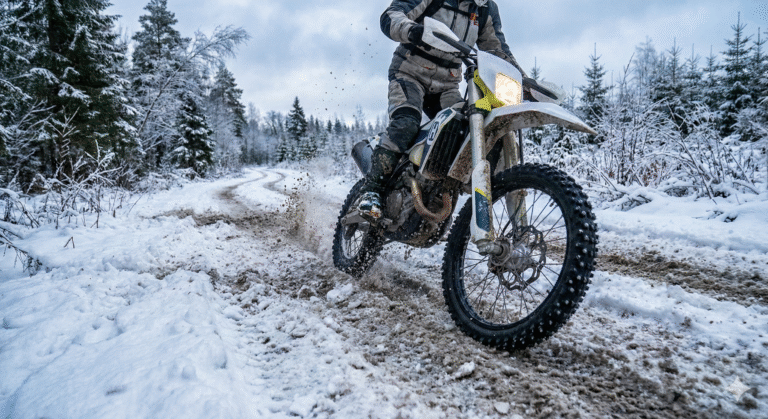 Motocyklista na motocyklu enduro wyposażonym w opony kolcowane, jadący dynamicznie przez błoto pośniegowe na leśnej drodze w zimowych warunkach.